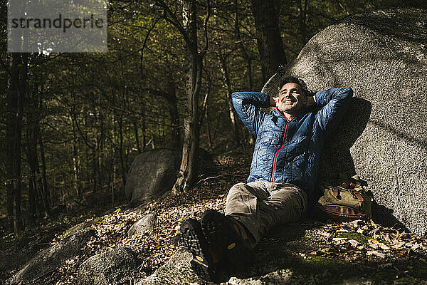 Glücklicher reifer Mann mit den Händen hinter dem Kopf  der sich an einem Felsen im Wald entspannt