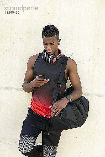 Junger Mann hält Sporttasche in der Hand und benutzt Smartphone  das an Wand lehnt