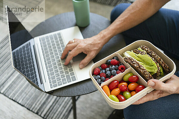 Die Hand eines Mannes hält eine Lunchbox mit gesundem Essen in der Hand und benutzt zu Hause einen Laptop