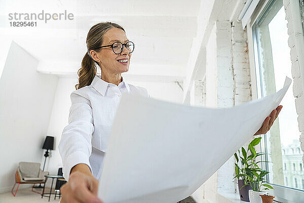 Glückliche Geschäftsfrau mit Brille blickt im Büro auf Blaupause