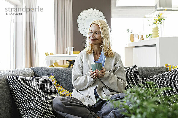 Lächelnde Frau mit Teetasse sitzt zu Hause auf dem Sofa