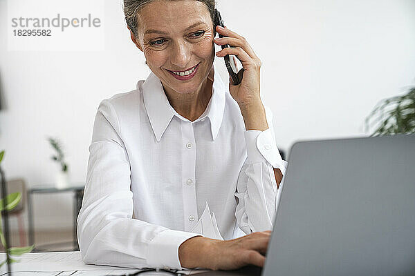 Glückliche reife Geschäftsfrau  die im Büro mit dem Laptop telefoniert