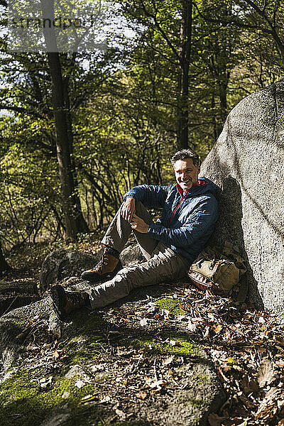 Lächelnder Mann mit Rucksack sitzt an einem Felsen im Wald