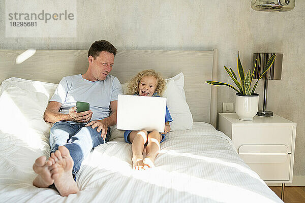 Lächelnder Mann mit Mobiltelefon schaut Tochter an  die Laptop im Bett benutzt