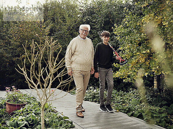 Großvater und Enkel gehen im Garten spazieren. Junge trägt Gartengabel
