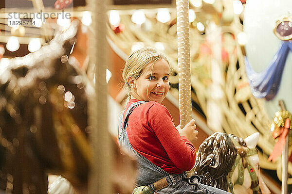 Lächelndes blondes Mädchen  das Spaß beim Sitzen auf einem Karussellpferd hat
