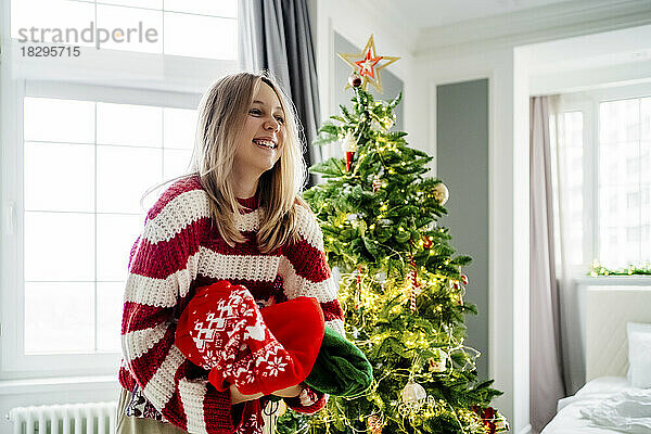Fröhliches Mädchen hält zu Hause Pullover am Weihnachtsbaum