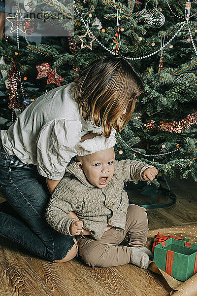 Schwester spielt mit ihrem kleinen Bruder vor dem Weihnachtsbaum