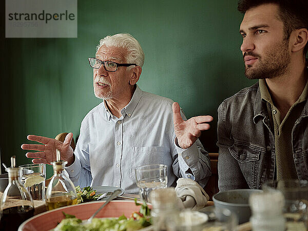 White-haired man sitting awith adukt son at lunch table gesturing
