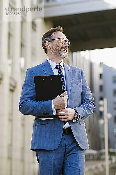 Happy mature businessman with clipboard standing in front of building
