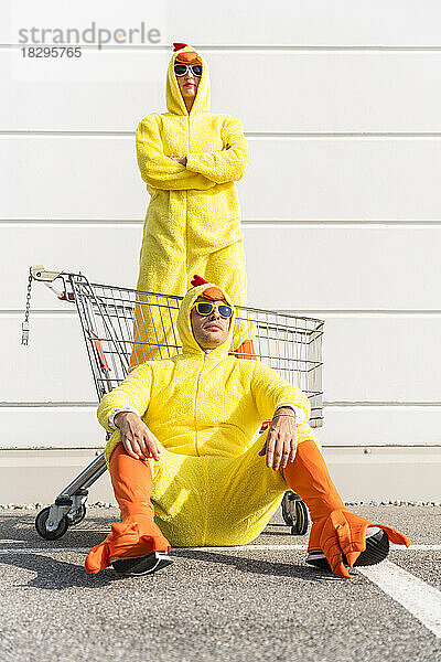 Frau im Hühnerkostüm steht im Einkaufswagen  Mann sitzt auf Fußweg