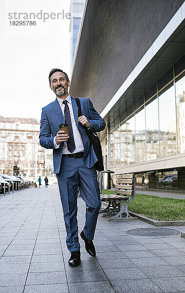Lächelnder reifer Geschäftsmann mit Einwegkaffeetasse  der auf dem Fußweg läuft