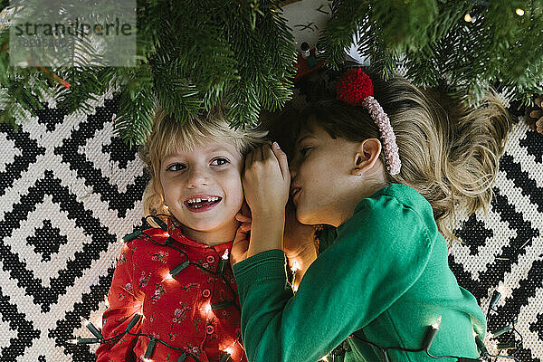Mädchen flüstert Schwester ins Ohr und liegt mit beleuchteten Lichterketten neben dem Weihnachtsbaum