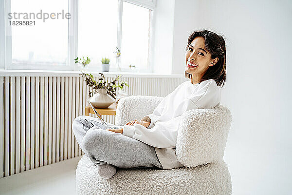 Glückliche junge Frau mit Laptop sitzt zu Hause auf einem Stuhl