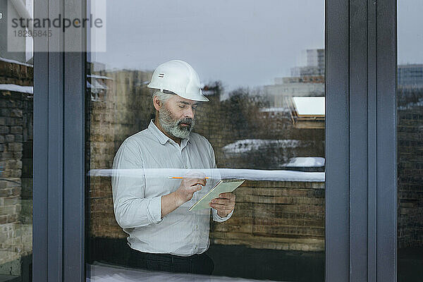 Geschäftsmann mit Schutzhelm macht sich Notizen auf der Baustelle  gesehen durch Glas