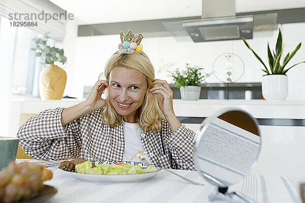 Glückliche Frau mit Kronenstirnband sitzt am Esstisch