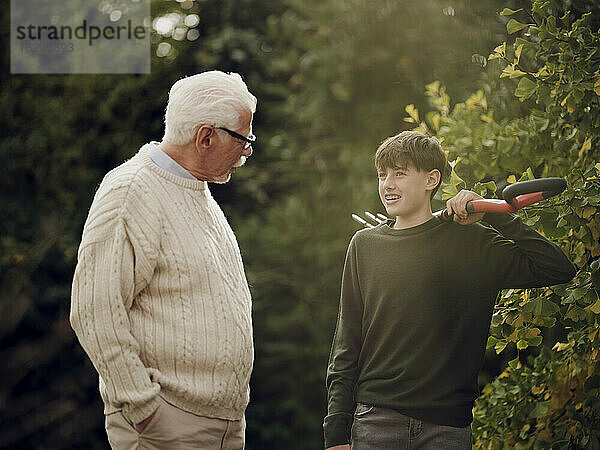 Großvater und Enkel unterhalten sich im Garten. Junge trägt Gartengabel