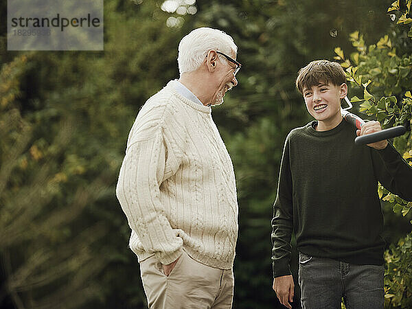 Großvater und Enkel unterhalten sich im Garten. Junge trägt Gartengabel