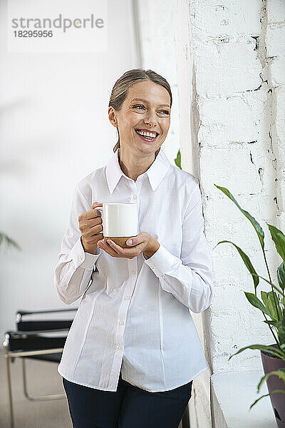 Glückliche reife Geschäftsfrau mit Kaffeetasse  die an der Wand steht