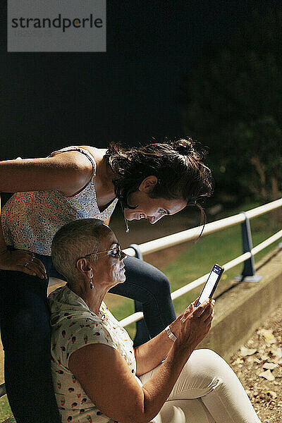 Ältere Frau teilt Mobiltelefon mit Freundin  die auf Geländer im Park sitzt