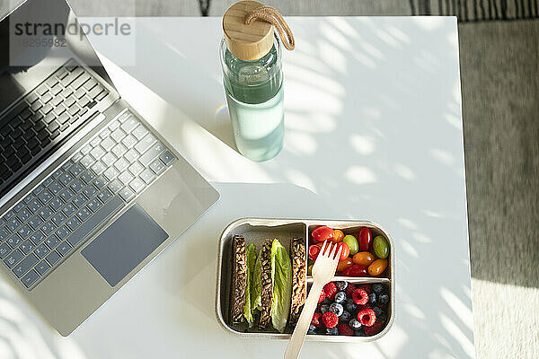 Roggenbrot  Beeren und Salat mit Einweggabel in der Lunchbox auf dem Tisch neben Wasserflasche und Laptop