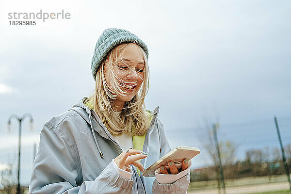 Lächelndes blondes Mädchen  das unter dem Himmel ein Mobiltelefon benutzt