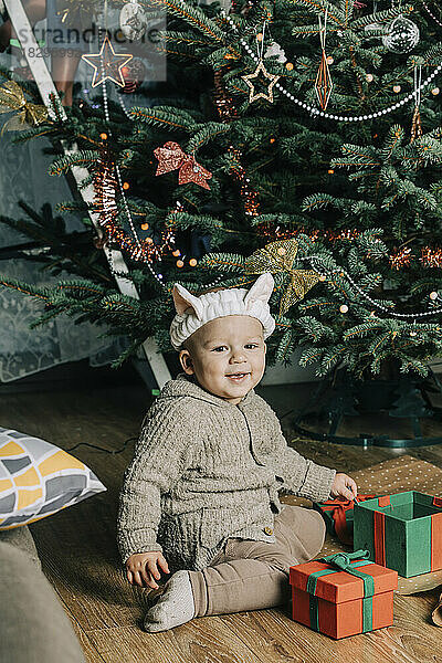Glücklicher kleiner Junge mit Geschenkboxen  der vor dem Weihnachtsbaum sitzt