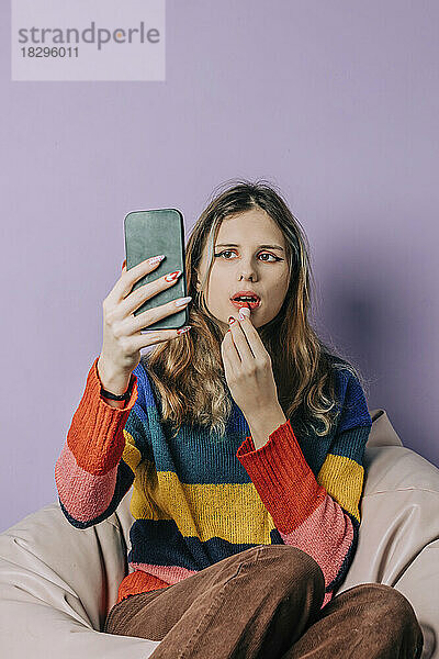 Mädchen blickt auf Smartphone und trägt Lippenstift vor violettem Hintergrund auf