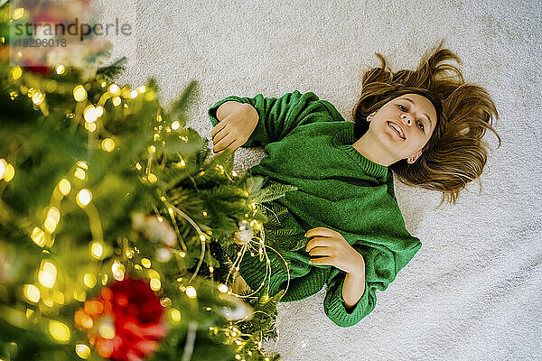 Glückliches Mädchen  das zu Hause auf einem Teppich unter einem beleuchteten Weihnachtsbaum liegt
