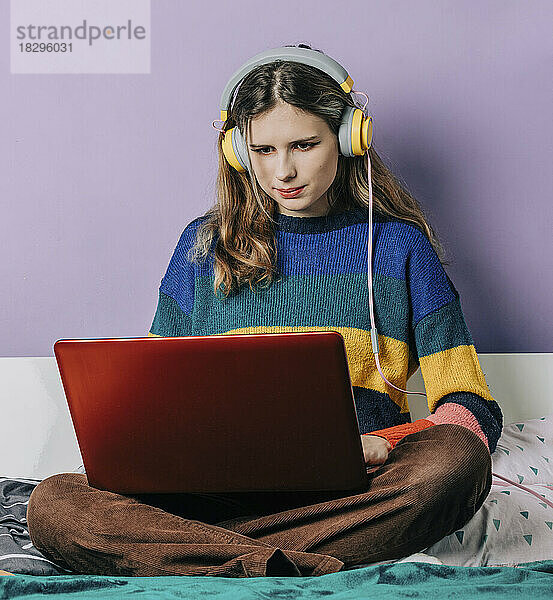 Mädchen mit Kopfhörern erledigt Hausaufgaben mit Laptop vor lila Wand
