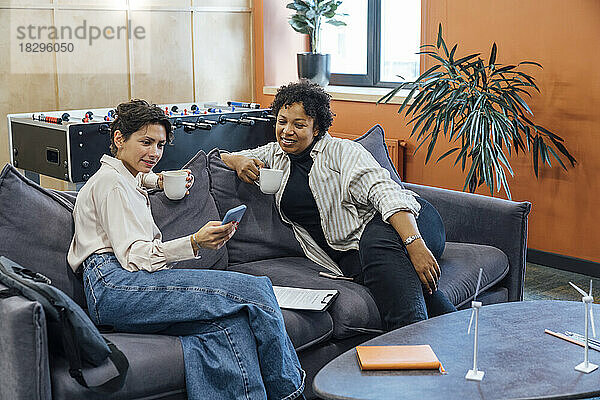 Smiling businesswoman with colleague using smart phone on sofa at office