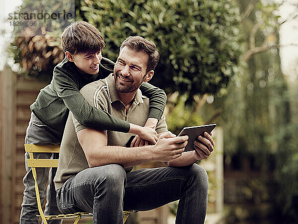 Son embraing father sitting in garden using digital tablet