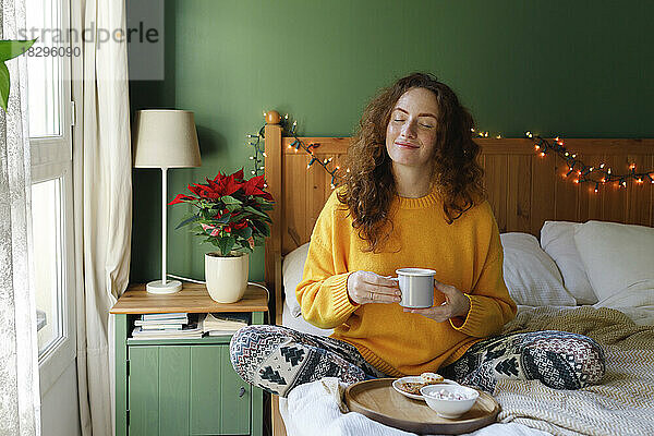 Frau mit geschlossenen Augen hält zu Hause eine Tasse Tee im Bett