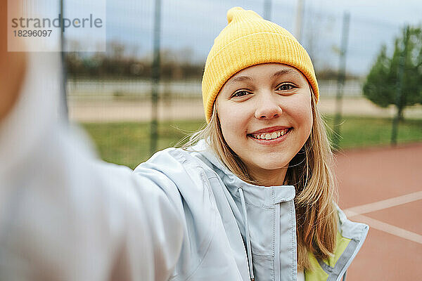 Fröhliches Teenager-Mädchen mit gelber Strickmütze macht ein Selfie auf dem Spielplatz