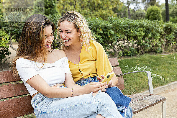 Glückliche Frau  die ihr Mobiltelefon mit einer Freundin auf einer Bank im Park teilt