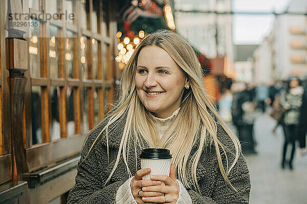 Glückliche blonde Frau mit Kaffeetasse steht auf Fußweg am Weihnachtsmarkt