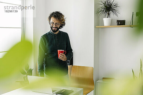 Glücklicher Freiberufler mit Kaffeetasse und Laptop am Schreibtisch im Büro