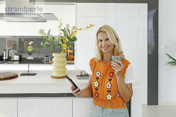 Lächelnde Frau mit Smartphone und Teetasse steht in der Küche