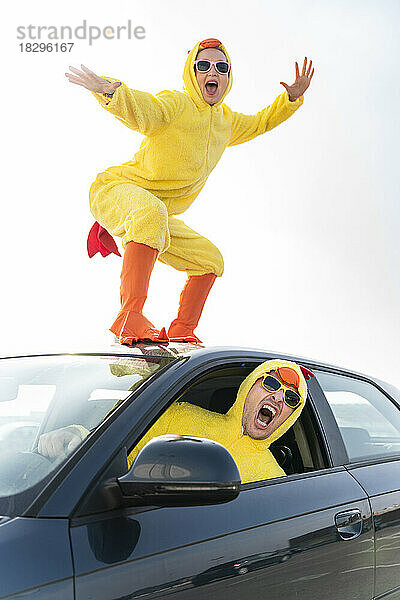 Mann fährt Auto mit unbeschwerter Frau im Hühnerkostüm  die auf dem Dach tanzt
