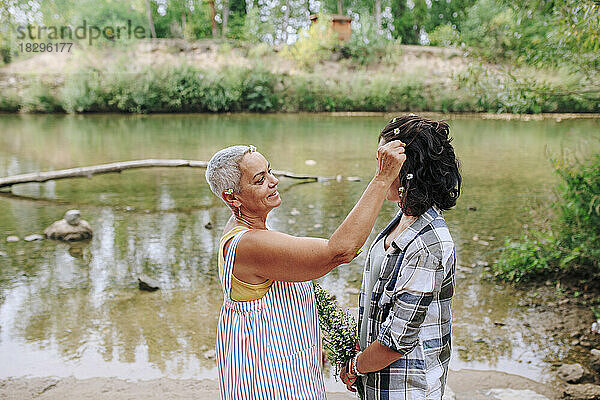 Ältere Frau schmückt die Haare einer Freundin mit Blumen am See im Park