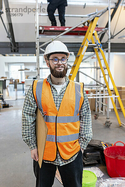 Lächelnder junger Ingenieur mit Klemmbrett auf der Baustelle