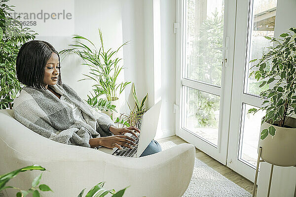 Junge Geschäftsfrau sitzt zu Hause im Sessel und benutzt Laptop