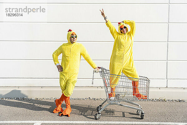 Mann und unbeschwerte Frau in Hühnerkostümen stehen mit Einkaufswagen vor der Wand