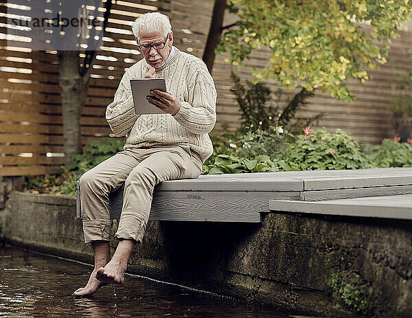 Älterer Mann sitzt am Gartenteich und nutzt digitales Tablet