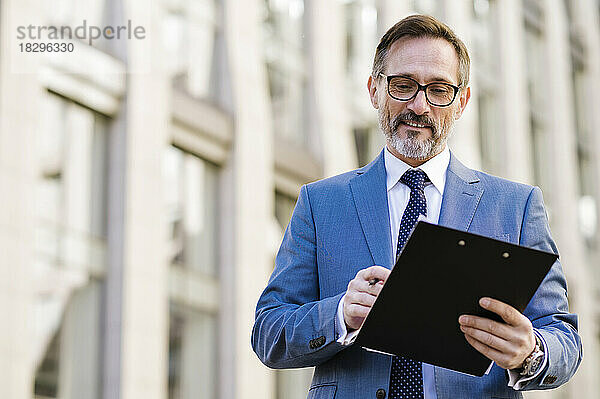 Smiling businessman with clipboard standing outside building
