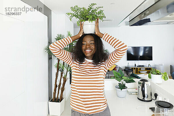 Glückliche Frau mit geschlossenen Augen balanciert Topfpflanze auf dem Kopf in der Küche