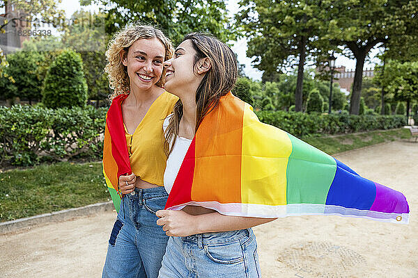 Lächelndes lesbisches junges Paar mit Regenbogenfahne bedeckt  das im Park spaziert