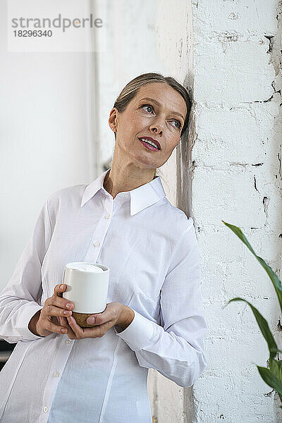 Nachdenkliche reife Geschäftsfrau mit Kaffeetasse lehnt an der Wand