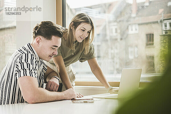 Lächelnde junge Geschäftsfrau diskutiert am Laptop mit Kollege am Schreibtisch