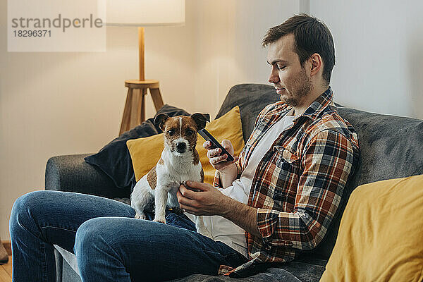 Mann benutzt Smartphone und sitzt mit Hund zu Hause auf dem Sofa